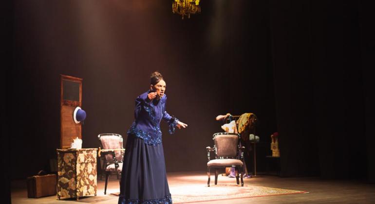 Atriz encena mãe de Sigmund Freud com vestido longo e azul em palco com cenário e com cadeira e chapeleiro antigo. 