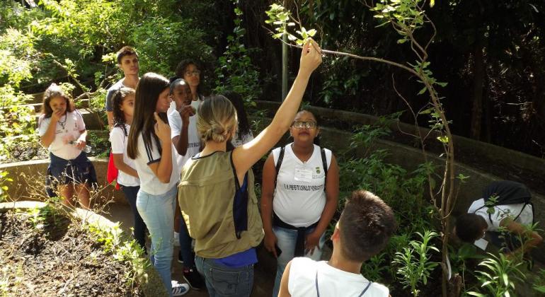 Monitora ambiental fala para grupo de jovens e adolescentes. A monitora mostra uma planta ao grupo