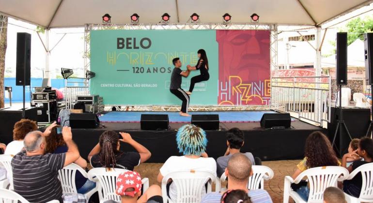 Dois jovens fazem acrobacias circenses no palco, assistidos cerca de cinco pessoas. 