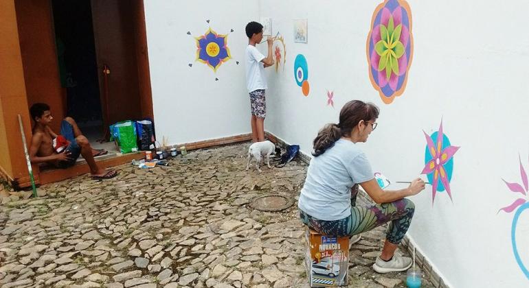 Criança sentada em na entrada de uma porta enquanto duas pessoas pintam mandalas coloridas em mural. 