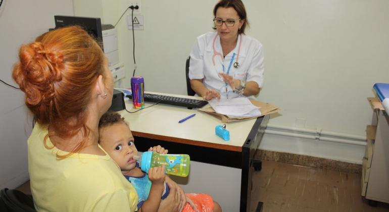 Mãe segura filhocom alergia a leite de vaca, que toma suco na mamadeira, ao fundo, atrás da mesa, médica realiza consulta. 