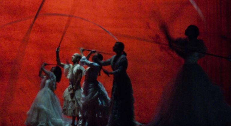 Proposta curatorial do FIT-BH 2018 tem o objetivo de horizontalizar e democratizar os processos relacionados ao evento. Foto: Ricardo Laf/PBH Em cena teatral, quatro mulheres de vestido longo movimentam astes, suas sobras são projetadas no fundo vermelho.