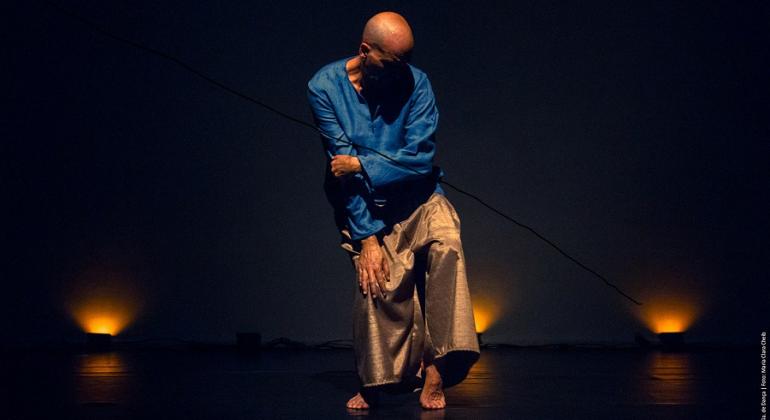 Homem careca vestido de azul roial e bege, faz misto de dança e performance em palco teatral com pouca iluminação. 