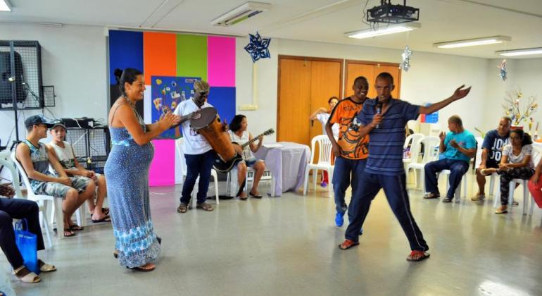 Mais de dez pessoas fazem parte de sarau com dança e violão em sala com cadeiras. 