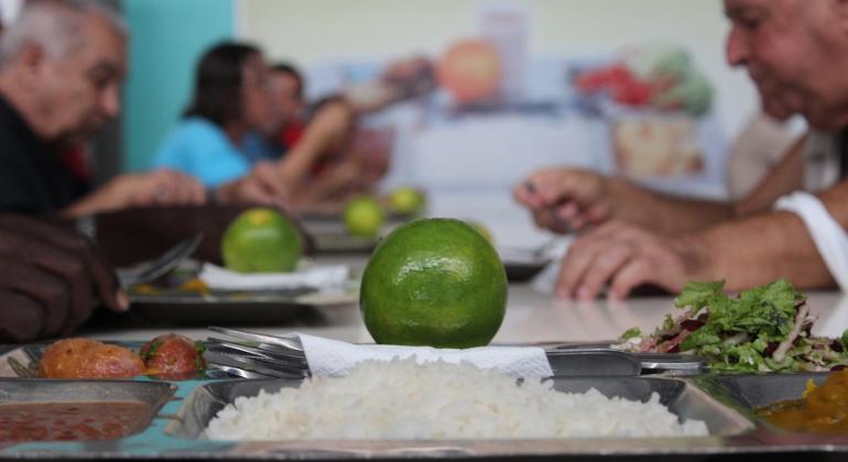 Bandejão de Restaurante popular com arroz, linguiça, salada, feijão e laranja; ao fundo pessoas fazem refeições. 