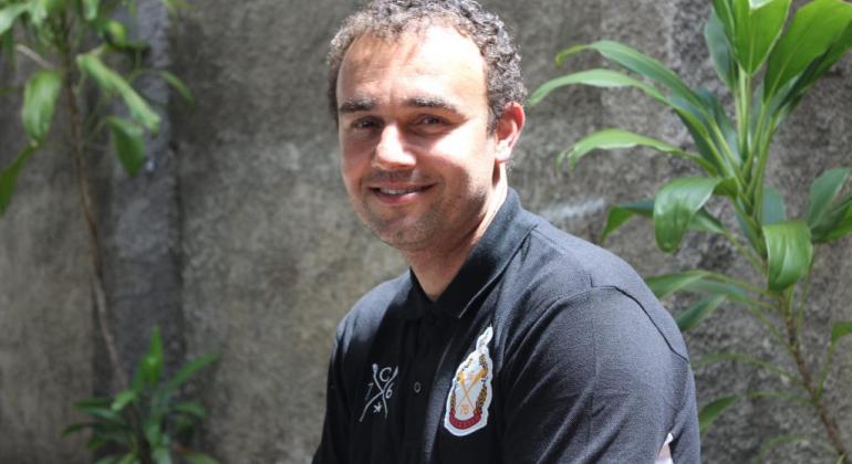 Acolhida, orientações e passagem para a cidade de origem são alguns dos serviços oferecidos no Serviço de Atenção ao Migrante da PBH. Foto: Débora de Oliveira Rapaz sorri, ao fundo, plantas e muro.