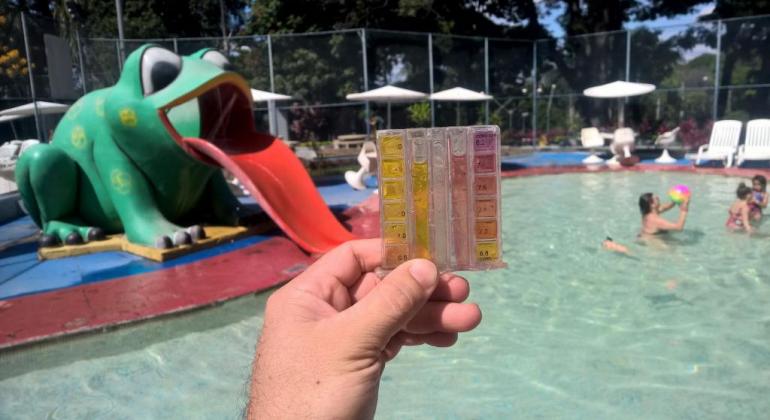 Mão de técnico da Vigilância Sanitéria com artefato plástico que verificaa qualidade da água; ao fundo, piscina com pessoas brincando dentro dela. 