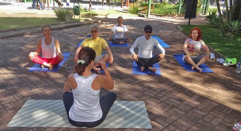 Na foto, seis pessoas estão sentadas no parque em posição de yoga