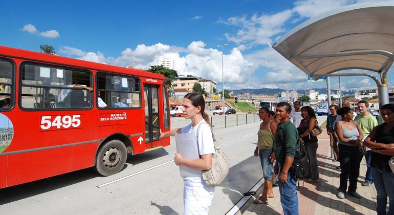 Sete cidadãos esperam no ponto de ônibus; à esquerda, parte de ônibus intermunicipal. 
