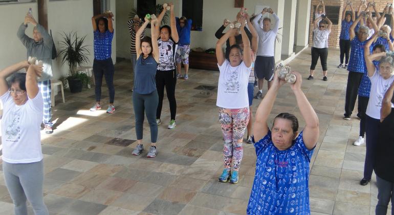 Lian Gong oferecido pela PBH auxilia no combate a dores articulares e musculares. Foto: Naara Dias/PBH Cerca de vinte pessoas, trajando roupas leves e flexíveis, erguendo pequenos pesos acima da cabeça.