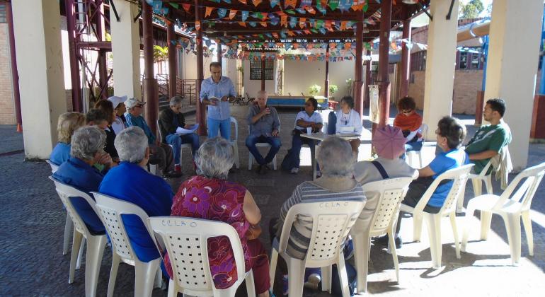 Cerca de quinze pessoas sentadas em círculo participam do Sarau. Um homem, de pé, lendo poesia detém a atenção de todos. 