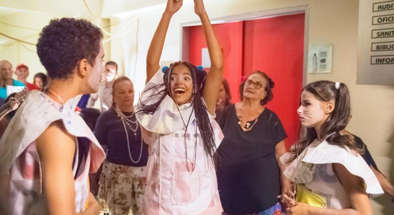 Alunos aprofundam conhecimento sobre teatro em Centro Cultural do Barreiro. Foto: Ricardo Laf Cerca de dez pessoas em um corredor observam três artistas, caracterizados para apresentação e com as faces artisticamente pintadas, fazendo movimentos teatrais.