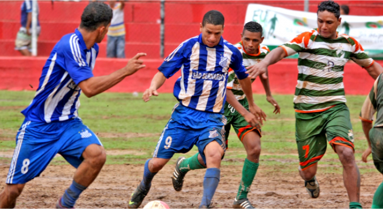 Quatro jogadores de futebol, dois de uniforme azul e dois de uniforme verde, disputam uma bola em uma partida da copa Centenário de Futebol Amador.