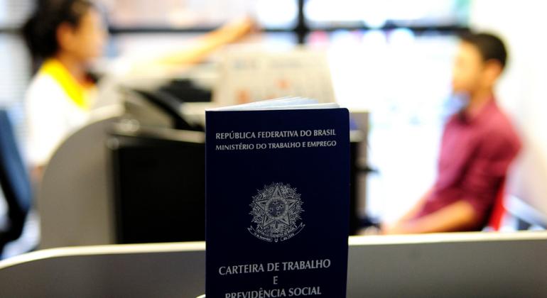 Carteira de trabalho à frente; ao fundo, homem é entrevistado por mulher. 