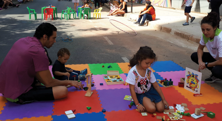 Duas crianças, supervisionadas por adulto, brincam de jogos infantis como jogo da memória em superfície colorida, ao fundo, na rua, mesas, cadeiras, e três adultos passeando