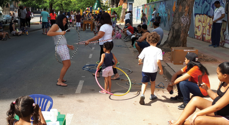 Quatro crianças brincam de bambolê em rua fechada, acompanhadas por jovens sentados da calçada; ao fundo, mais pessoas passeiam. 