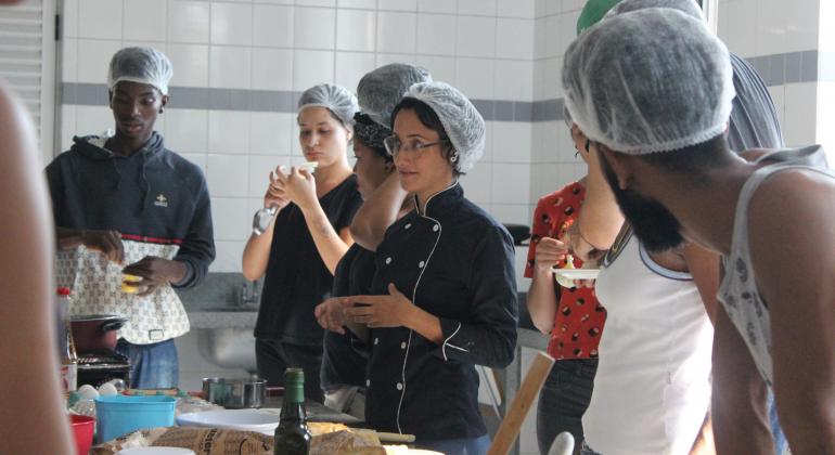 Cerca de seis alunos, com touca de proteção higiênica, assistem aula de gastronomia. 