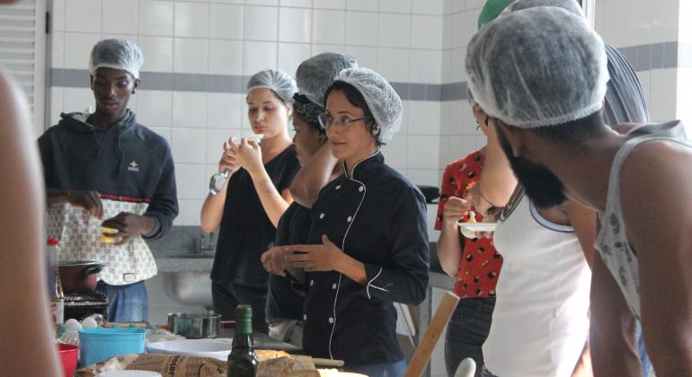 Gastronomia, de artes visuais, de teatro e de arte circense são as oficinas oferecidas por parceria da PBH com a UFMG. Foto: Débora de Oliveira/PBH Cerca de seis alunos, com touca de proteção higiênica, assistem aula de gastronomia.