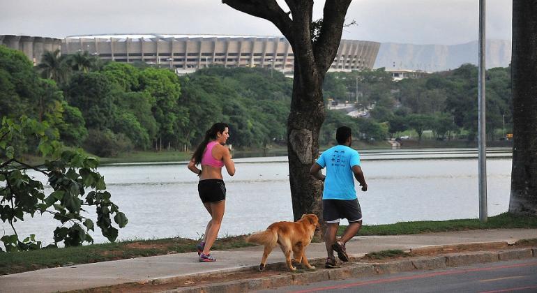 Duas pessoas caminham na orla da Lagoa da Pampulha com um cão. 