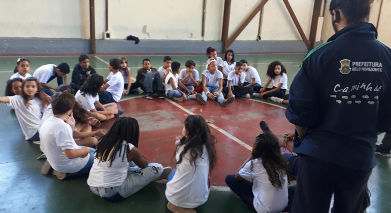 Cerca de vinte alunos sentados em círculo e um professor de pé, trajando uma blusa do programa Caminhar.