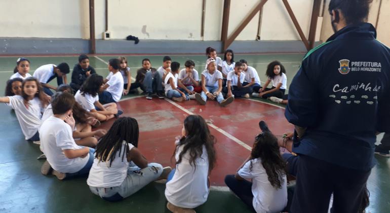 Caminhar na Escola oferece atividades físicas a estudantes da rede municipal. Foto: Vilma Tomaz/PBH Cerca de vinte alunos sentados em círculo e um professor de pé, trajando uma blusa do programa Caminhar.