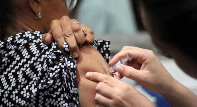 Campanha de vacinação contra a Influenza vai até 1º/6 Idosa de costas puxa a blusa preta enquanto é vacinada por agente de saúde