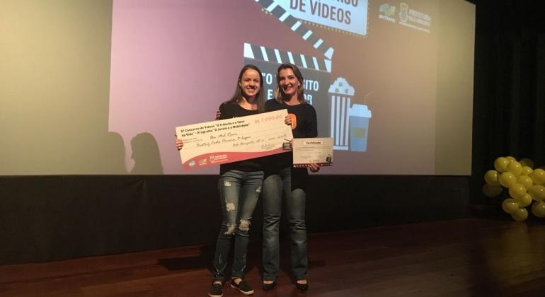 Duas vencedoras do 5ª edição do concurso de vídeos “O Trânsito e o Valor da Vida mostram o cheque recebido e sorriem. 