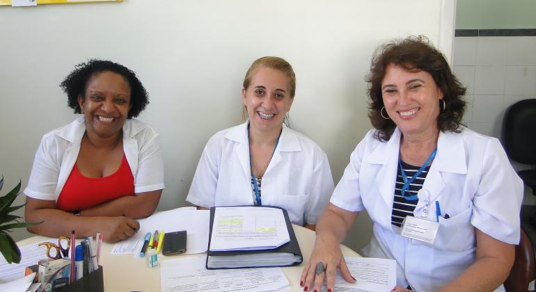 Centros de Saúde da região leste incentivam e promovem o aleitamento materno. Foto: Divulgação/PBH Três mulheres profissionais da saúde sentadas em uma mesa, onde há várias folhas e fichas.
