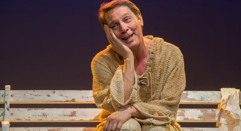 Peça teatral “Francisco de Assis – do Rio ao Riso” está em cartaz entre os dias 25 e 27/5, sexta e sábado, às 20h, e domingo, às 19h. Foto: Glaucia Rodrigues Ator Carlos Nunes sentado em banco de madeira, com a mão direita sobre o rosto, sorrindo, em apresentação teatral.