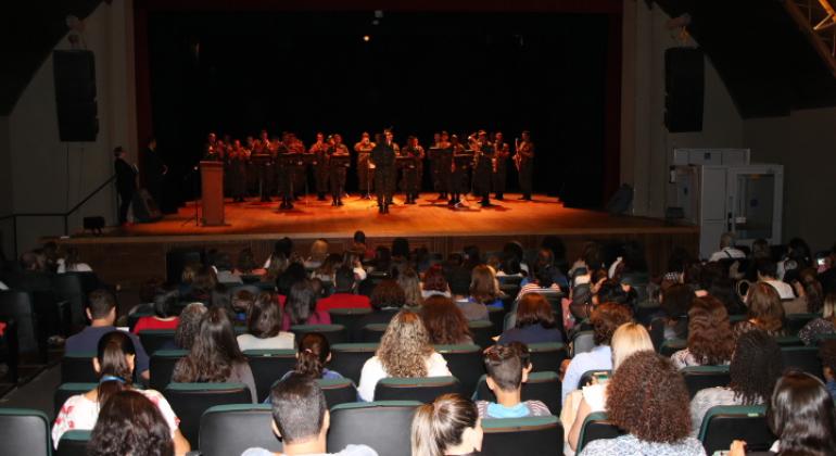 Auditório do Teatro Francisco Nunes com todas as cadeiras preenchidas por alunos, enquanto a Banda da Guarda Municipal se apresenta no palco