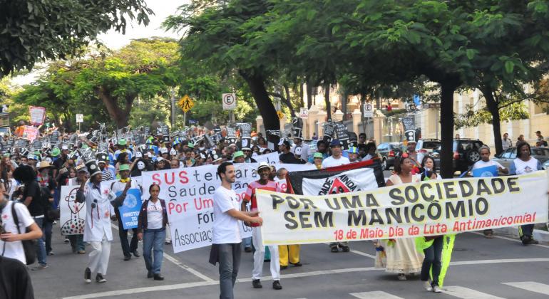 Mais de duzentas pessoas fazem passeata em avenida em prol da Luta Manicomial, com faixas. A faixa à frente da passeata tem os dizeres: "Por uma Sociedade sem Manicômio". 