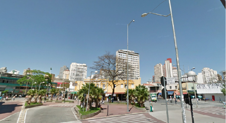Avenida Paraná, no Centro de Belo Horizonte, com céu azul, dois prédios altos no centro da foto. Há dois postes de energia e algumas plantas no canteiro central