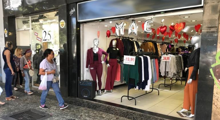 Com a proximidade do Dia das Mães, o Procon-BH vem realizando ações de fiscalização no comércio de rua e nos shoppings da cidade. Foto: Mônica Ramos/PBH Loja de roupas femininas da área central de BH com balões vermelhos em forma de coração e balões que formam a palavra "Mãe" em prateado; na calçada, passam três cidadãos.