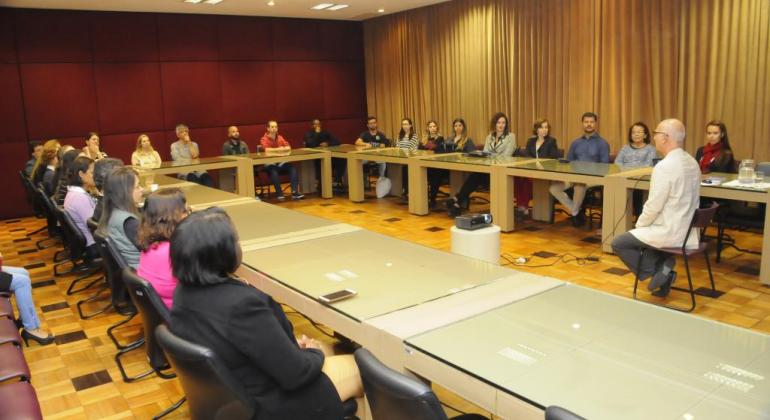Júlio Machado, consultor de desenvolvimento humano, sentado em uma cadeira, defronte a uma mesa em formato de "u" com cerca de 18 ouvintes. 