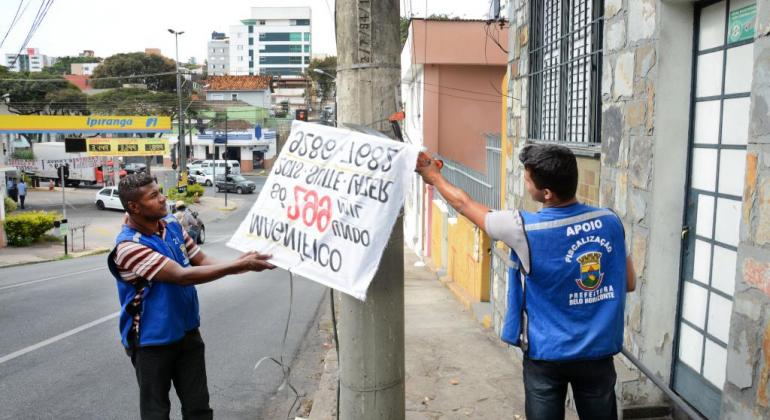 Dois fiscais da PBH retirando faixa irregular de um poste.