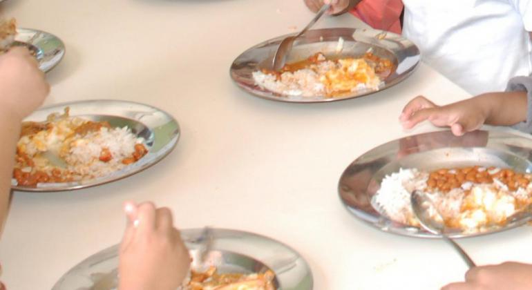Mesa com seis pratos onde estudantes se alimentam.