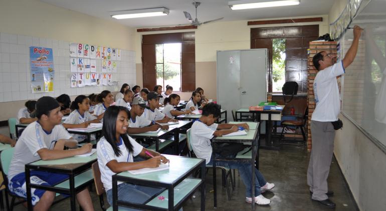 Professor escreve na lousa. Há cerca de 20 alunos sentados