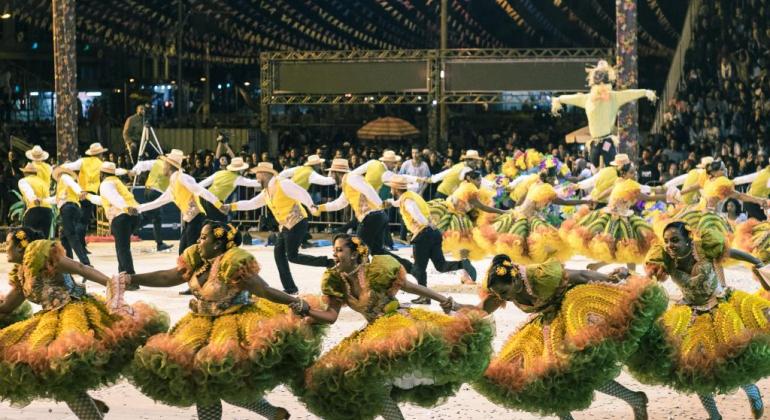 Mais de vinte membros de quadrilha junina fazem apresentação, vestidos de roupas juninas nas cores verde e amarelo. 