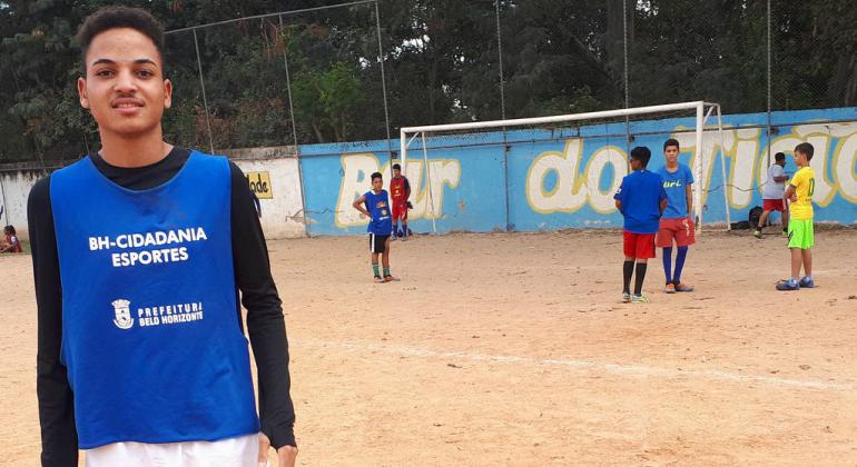 Esporte Esperança revela bons atletas Jovem posa para foto em campo de futebol de terra. O fundo tem outros colegas jogadores e a área do gol.