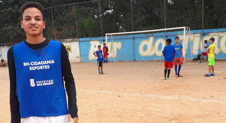 Jovem posa para foto em campo de futebol de terra. O fundo tem outros colegas jogadores e a área do gol.