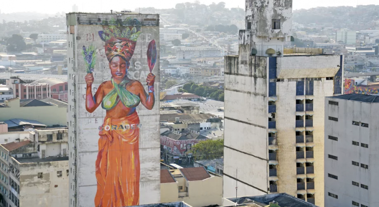 Mulher de origem africana, com turbante na cabeça e roupas em cores vibrantes, pintada em lateral de arranha-céu de Belo Horizonte, durante o dia. 