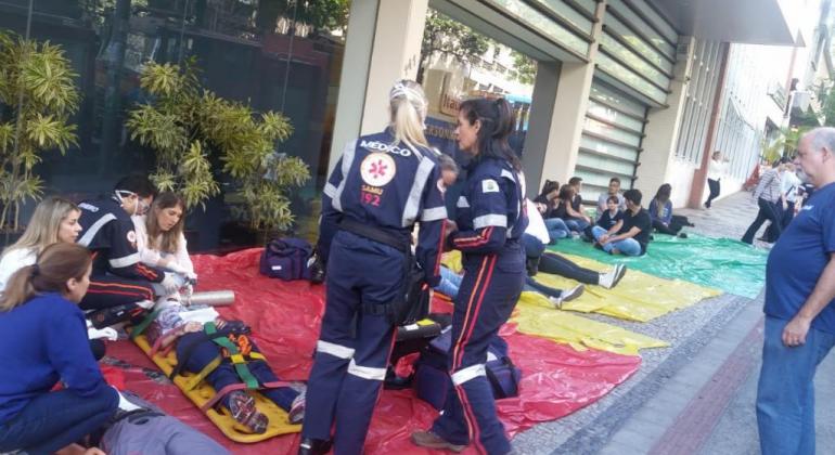 TRês membros da equipe do SAMU atendem a mais de dez pessoas deitadas no chão da calçada, simulando passar mal devido a incêndio simulado. 