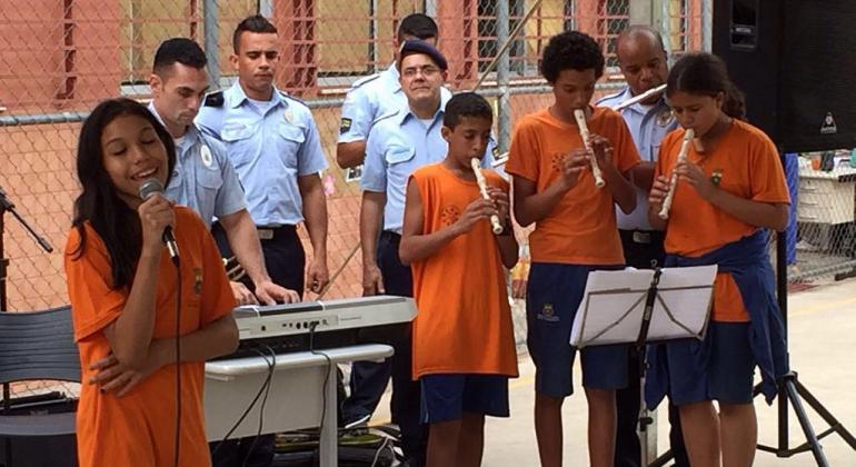 Três crianças tocam flauta, uma canta ao microfone e um guarda municipal toca treclado