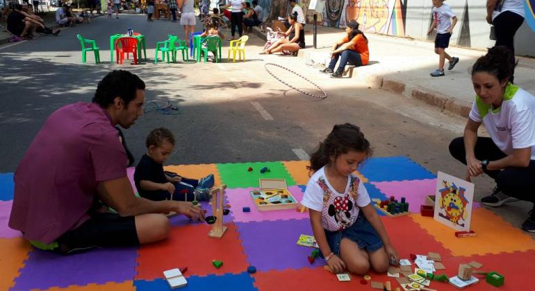 Duas crianças e dois adultos estão sentados em tapete colorido infantil em rua e brincam