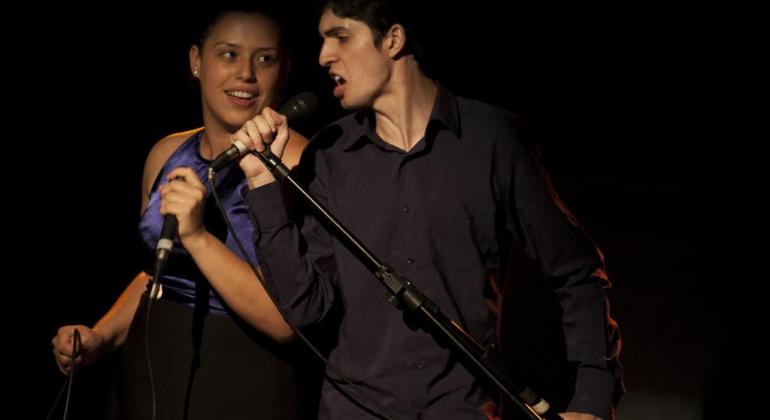 Dois jovens, um homem e uma mulher, cantam com microfones, em cena teatral de fundo escuro.