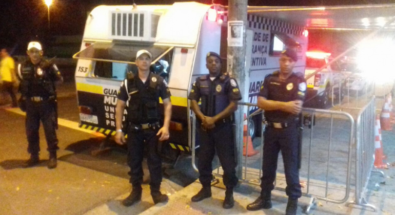 Quatro guardas municipais em frente a trailes da Guarda Municipal durante a noite. 