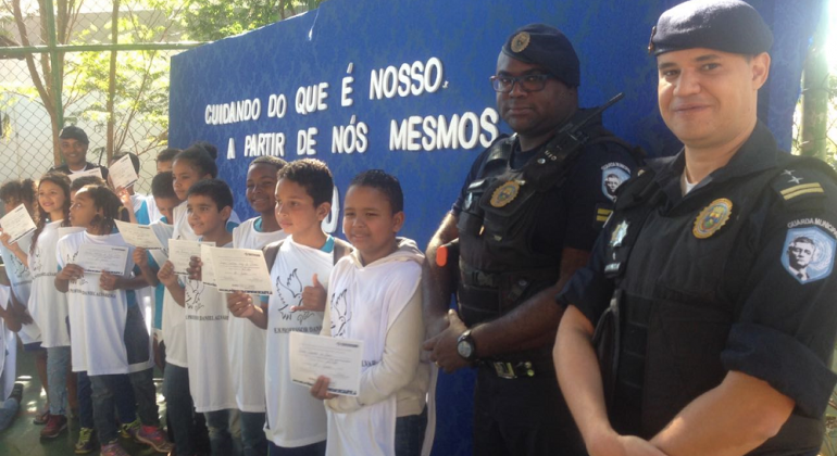 Alunos que debateram violência escolar recebem certificados de Guarda Municipal Dois guardas municipais e alunos das escolas municipais posam para foto com certificado.