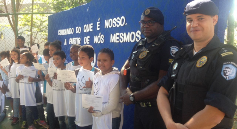 Dois guardas municipais e alunos das escolas municipais posam para foto com certificado.