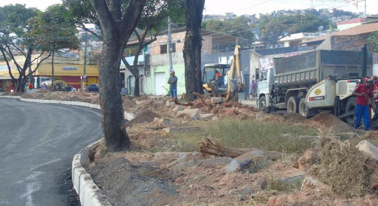 Caminhões e dpos funcionários ao lado de avenida com árvores, meio-fio recém pintado de branco e recapeamento novo. 