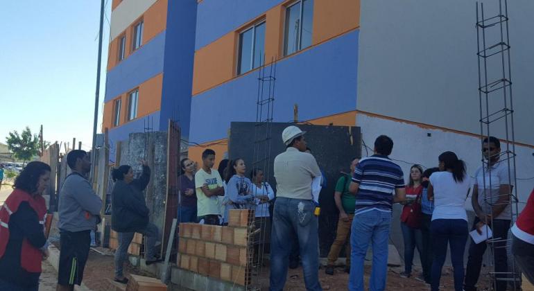 Cerca de dez pessoas, uma delas com capacete, prédio azul e laranja, cuja construção foi recém concluída, durante o dia.